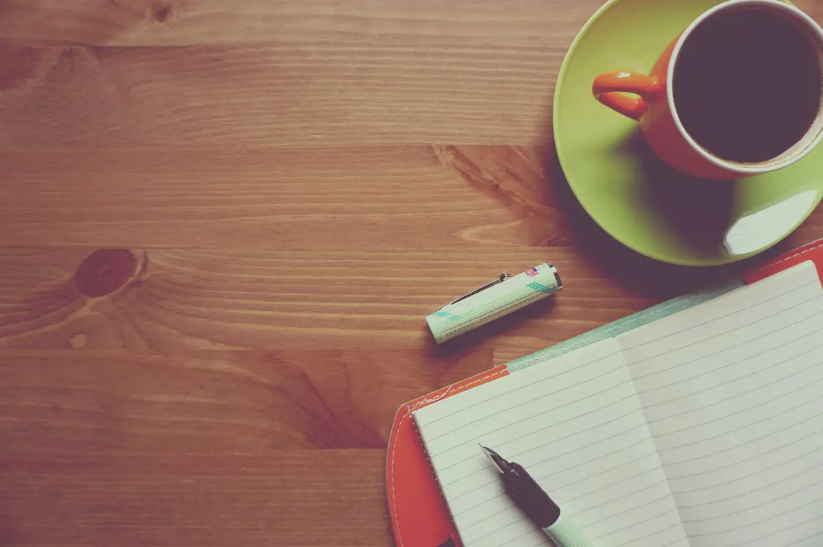 Caderno aberto com caneta e xícara de café sobre mesa de madeira