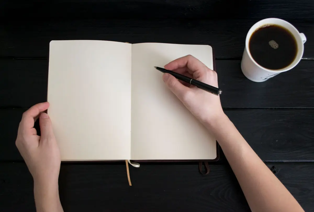 Mão com caneta sobre caderno em branco e xícara de café
