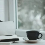 Caderno e xícara de café perto da janela, momento do diário estoico