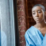 Paciente com roupa de hospital olhando pela janela, pensativa