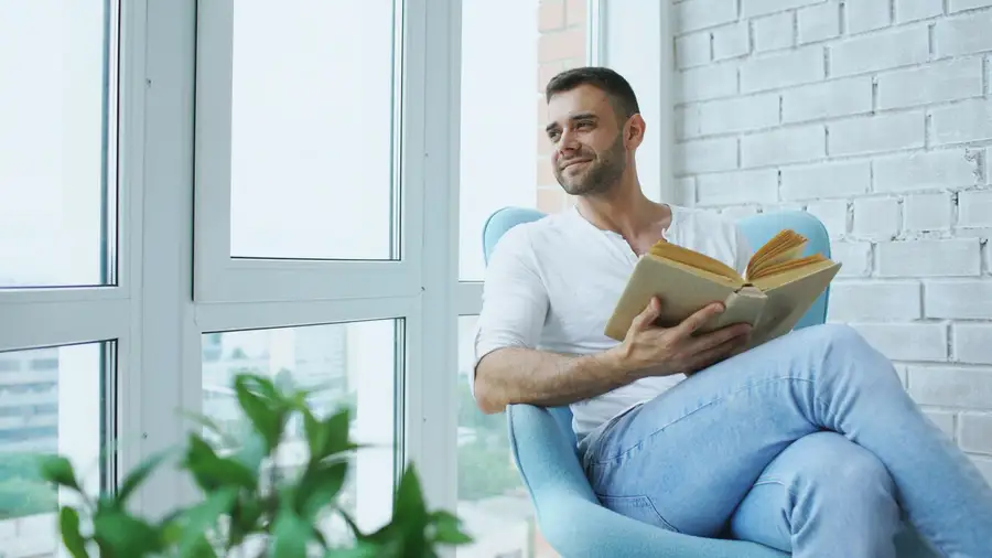 Homem sorrindo enquanto lê um livro sentado junto à janela