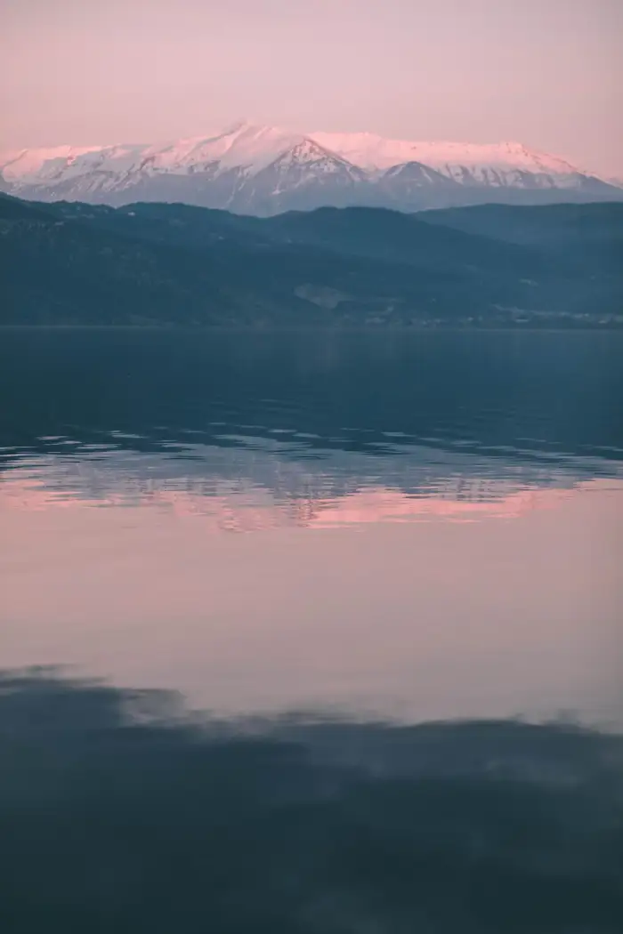 Lago calmo ao pôr do sol, com montanhas nevadas ao fundo