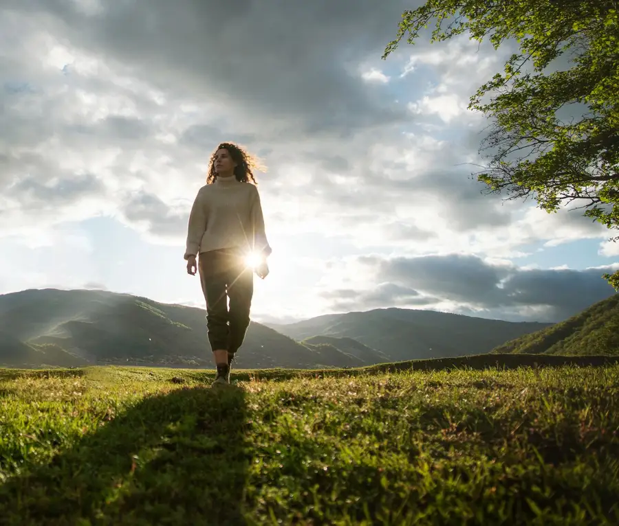 Mulher caminhando sozinha num campo verde ao sol, com montanhas ao fundo