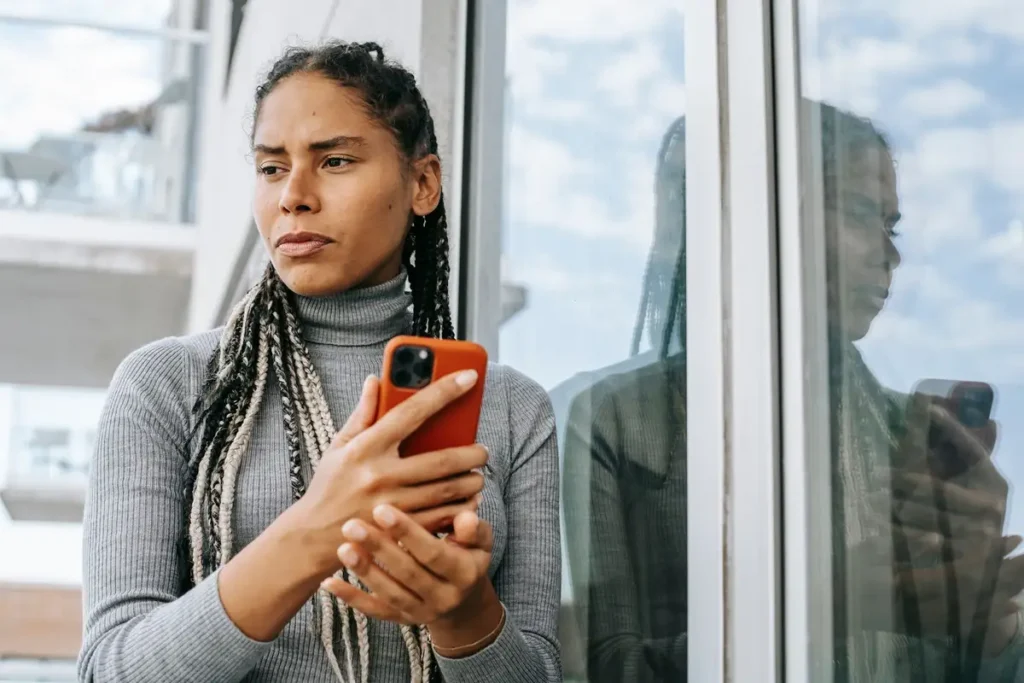 Mulher pensativa segurando o celular perto da janela