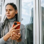 Mulher pensativa segurando o celular perto da janela